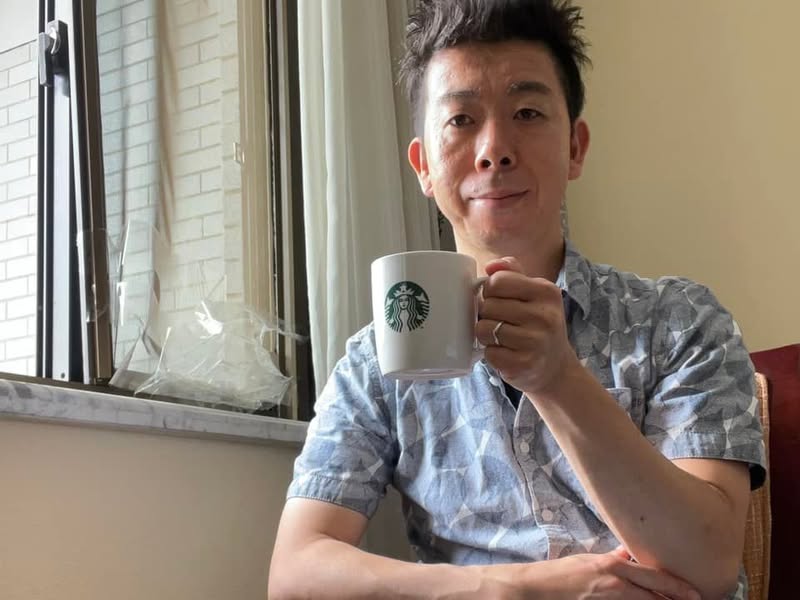 隔離ホテルの窓辺でコーヒーを持つ佐藤大介