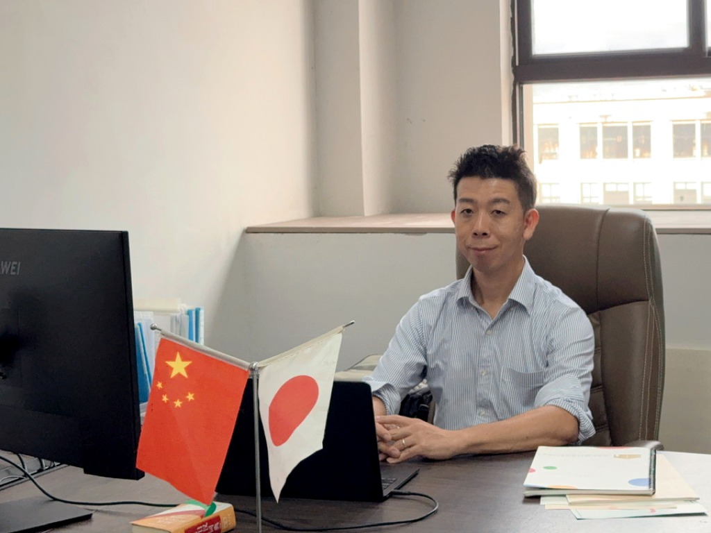 中国と日本の国旗が置かれた中国のオフィスでデスクに座る佐藤大介