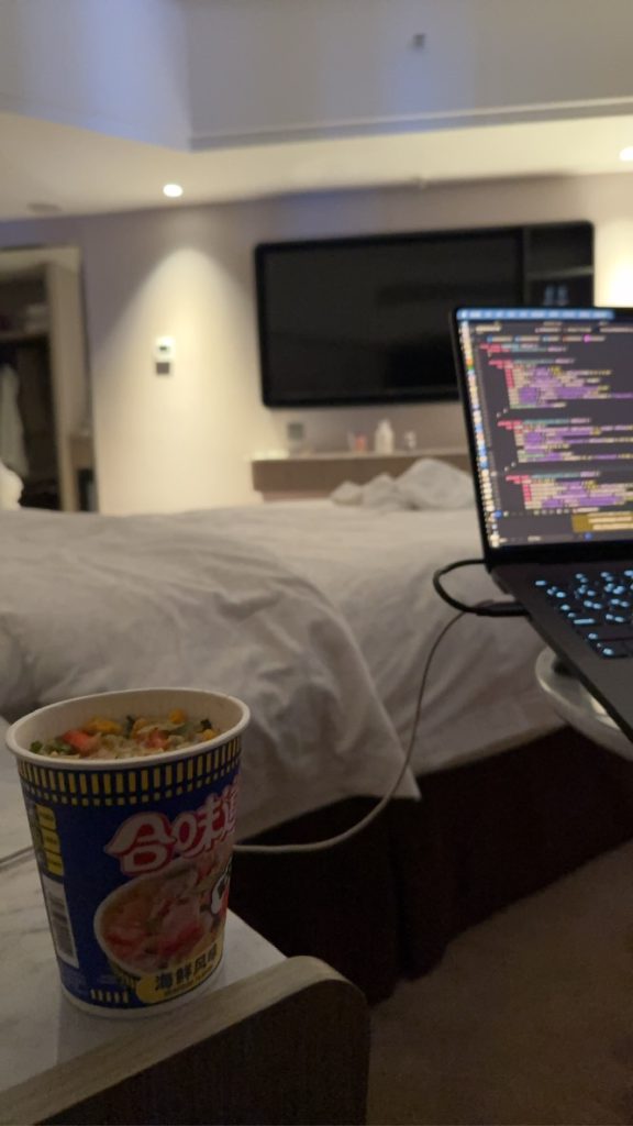 ホテルのベッド脇でカップ麺を置きノートパソコンのコードを確認している様子
